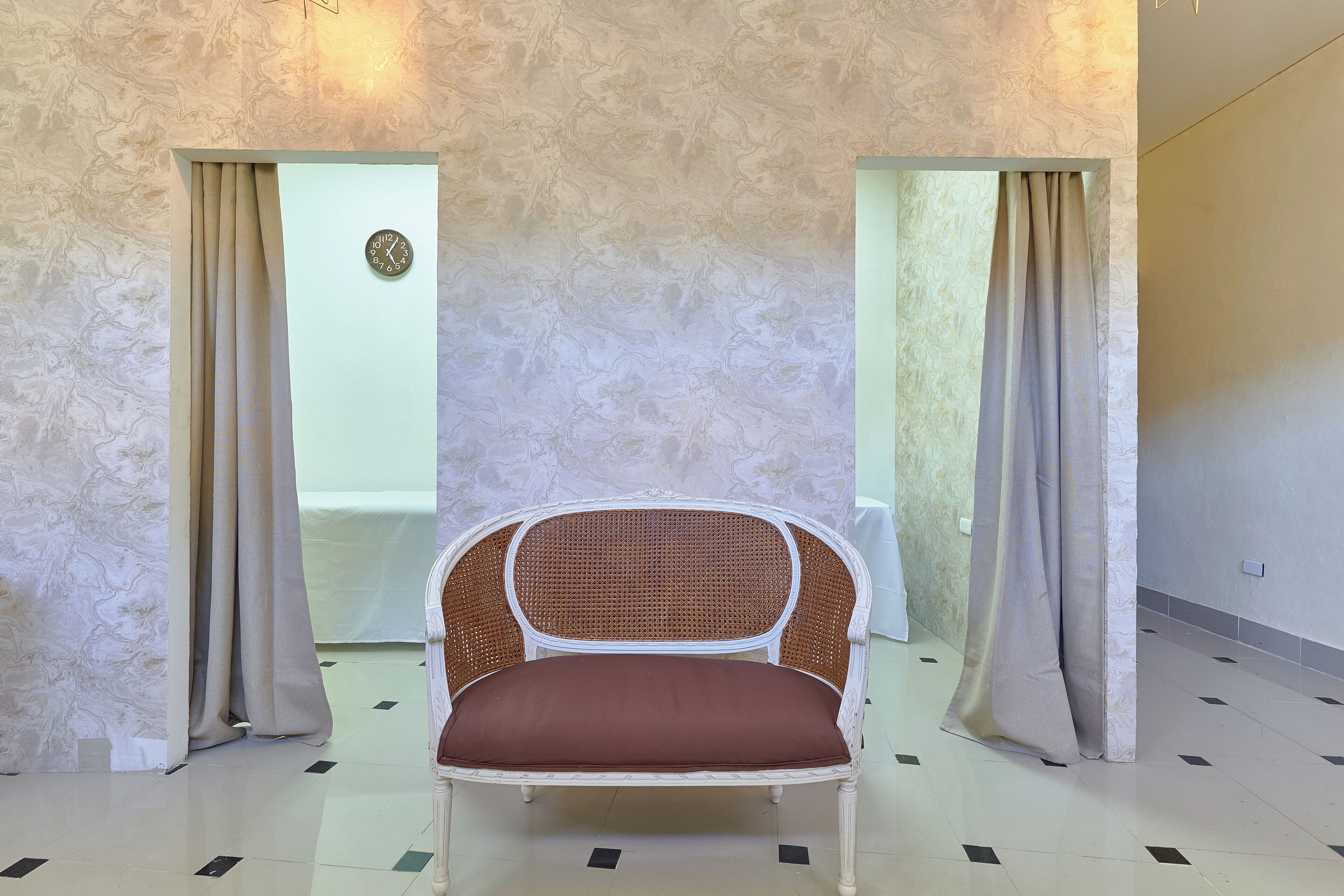 A room with a rattan chair, beige curtains, a clock on the wall, and a patterned floor.