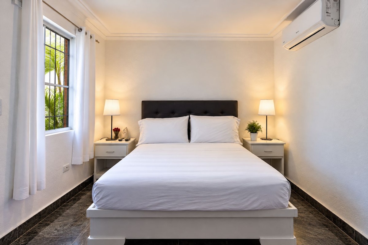 A hotel room with a large bed, two bedside tables, a window with curtains, and an air conditioning unit on the wall.