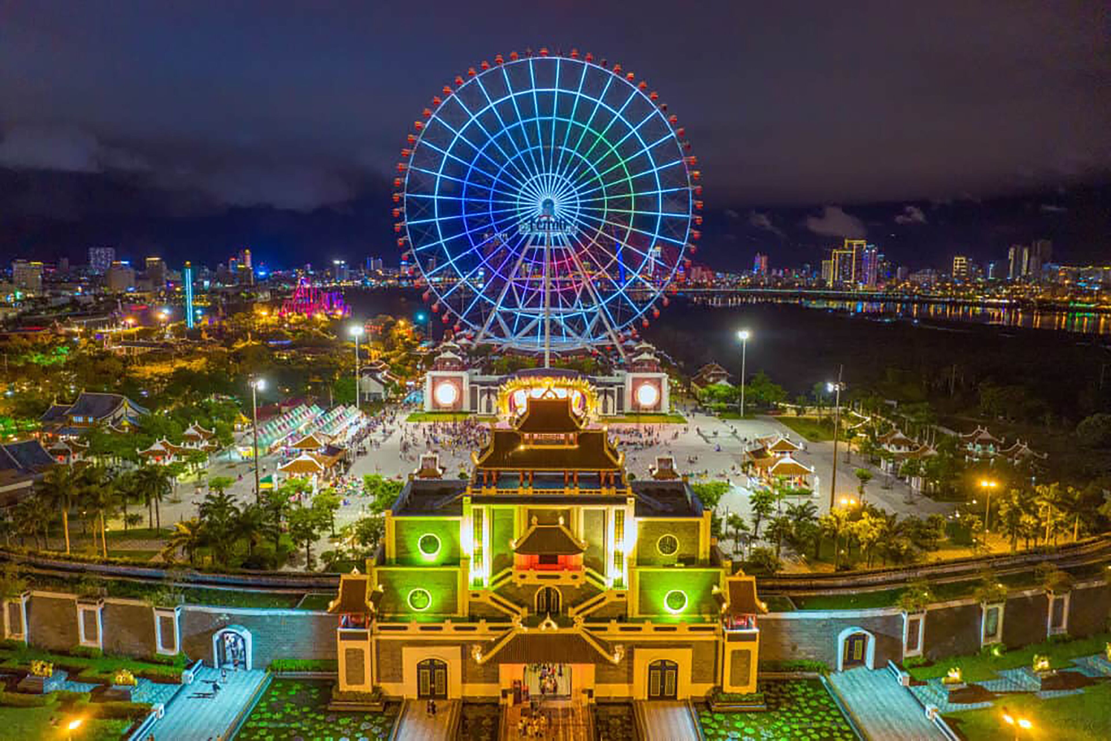 A large Ferris wheel with a colorful light display, a traditional building with green lights, and a waterfront area with buildings and a bridge in the background.