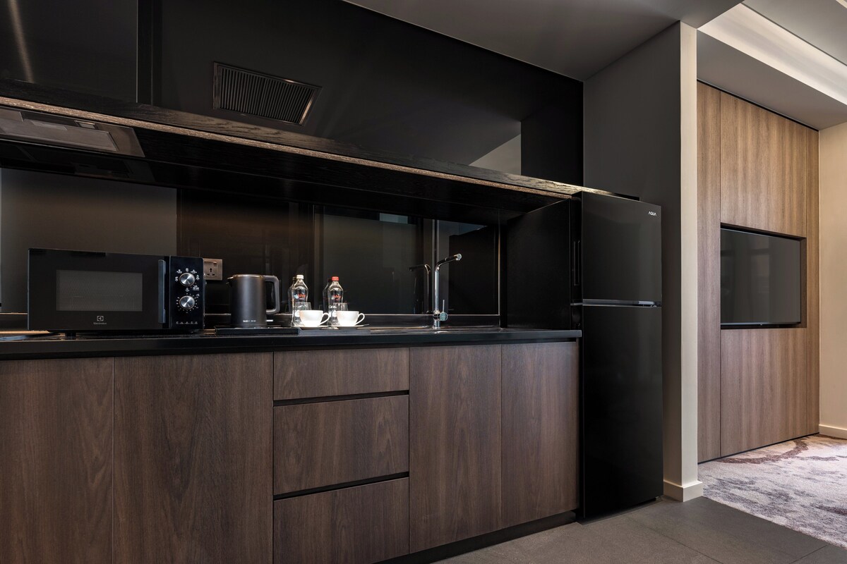 Una cocina moderna con armarios de madera oscura, encimera negra, microondas, hervidor y refrigerador.