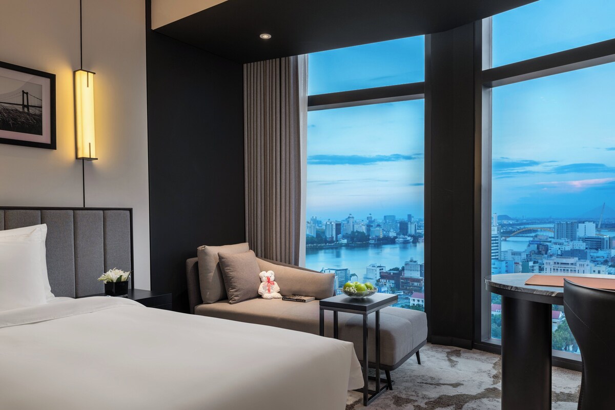 Habitación de hotel con cama, sofá, una mesita con frutero, un osito de peluche y vistas a la ciudad y al río.
