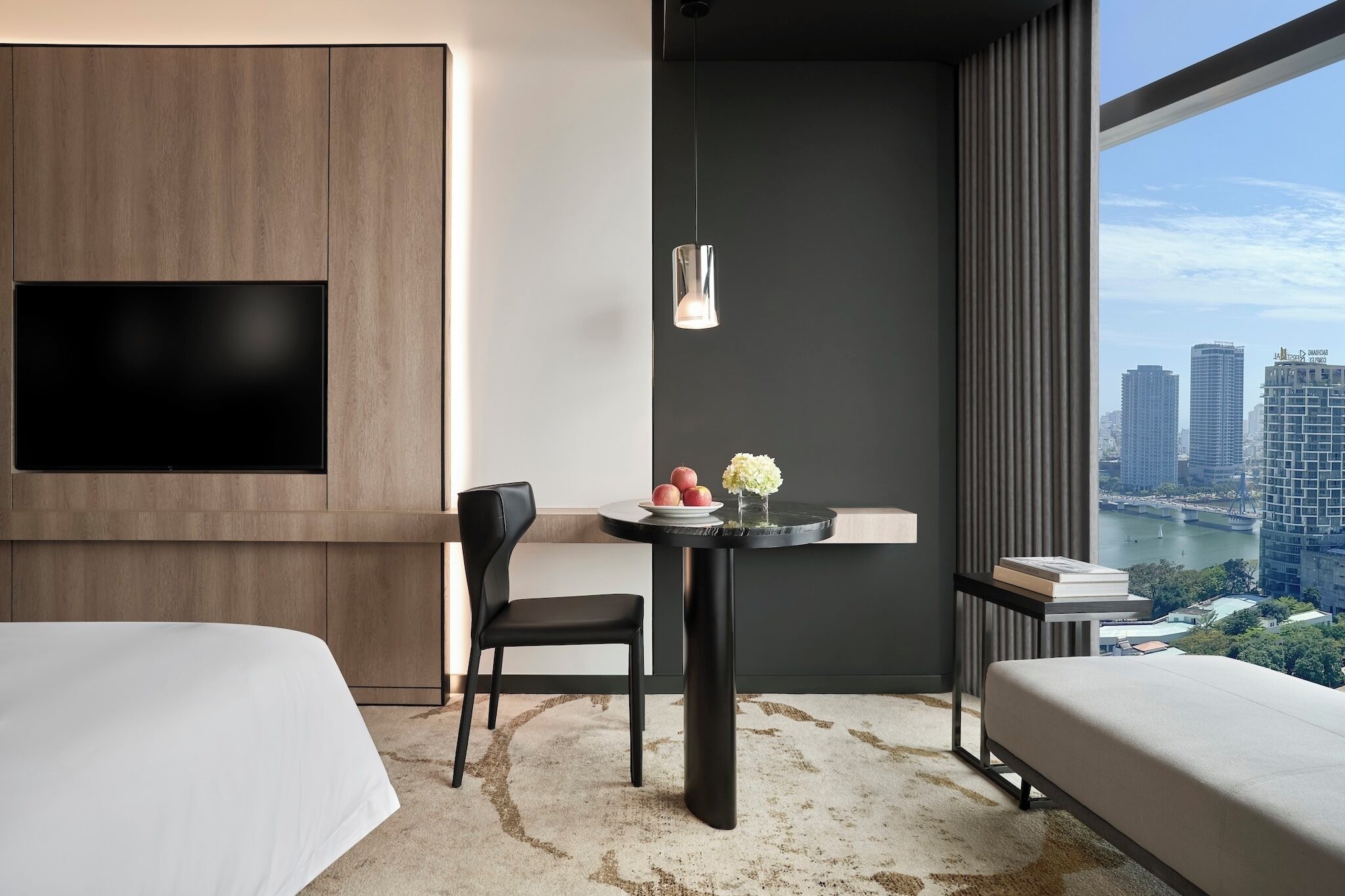 A modern hotel room with a large TV, a round table with a vase and fruit, a bed, and a view of the cityscape.