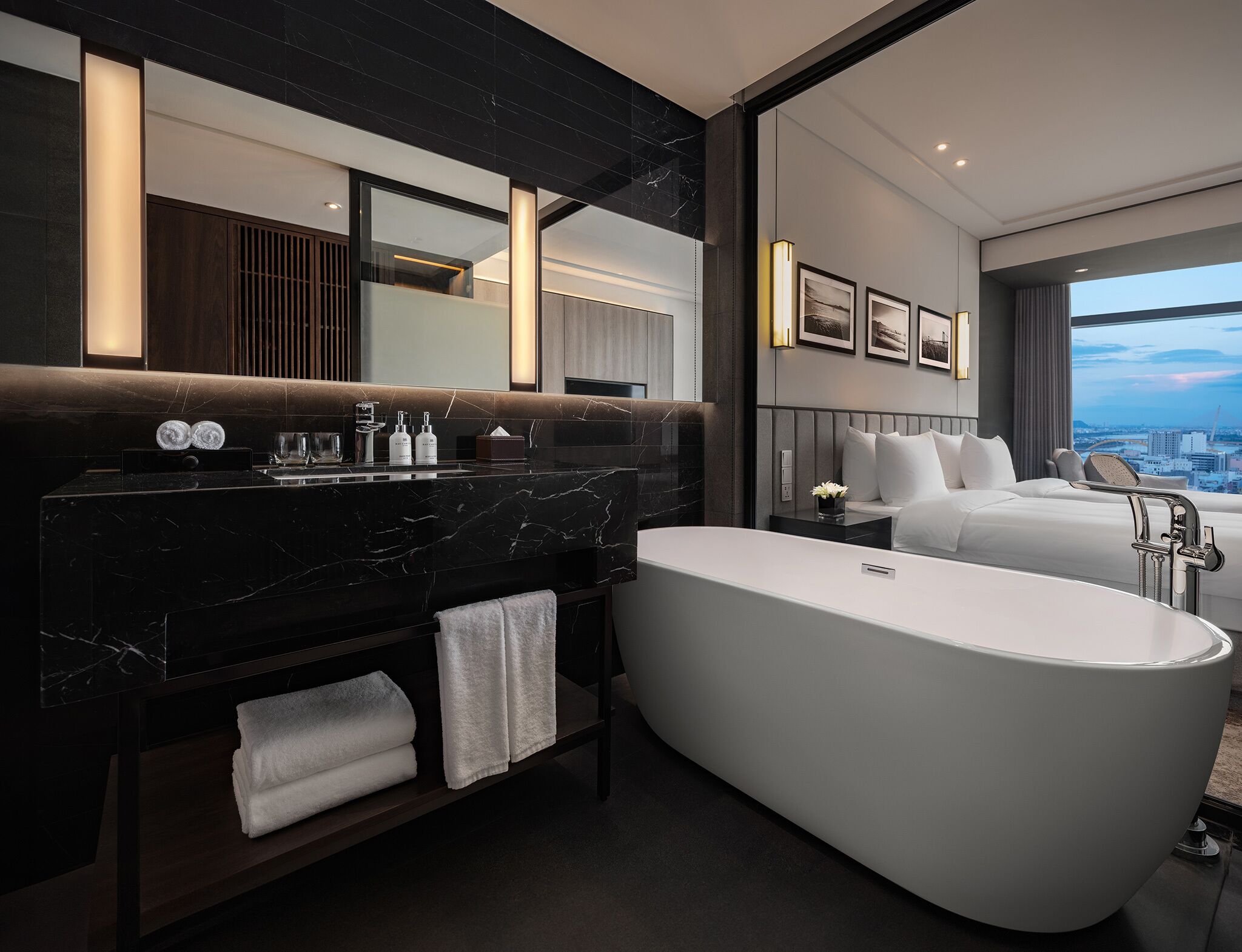 Une salle de bain d’hôtel moderne, dotée d’une grande baignoire, d’un meuble-lavabo avec un plan de travail en marbre foncé et d’un grand miroir. On y trouve un lit avec une literie blanche et une vue sur la ville par la fenêtre.