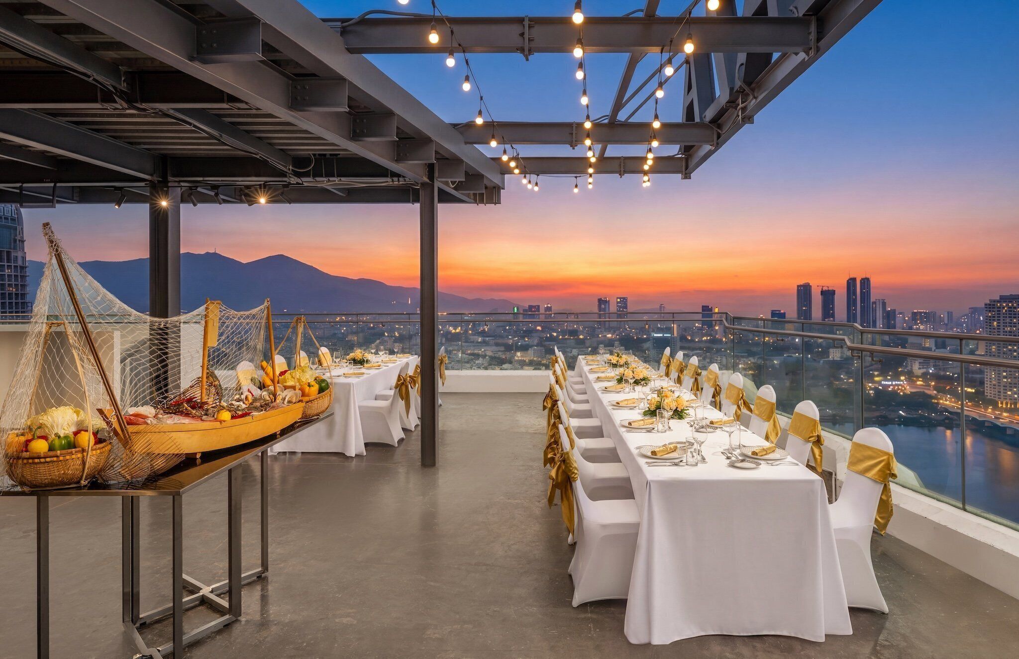 Un área de comedor en la azotea con mesas dispuestas para una comida, con vistas a un paisaje urbano al atardecer.