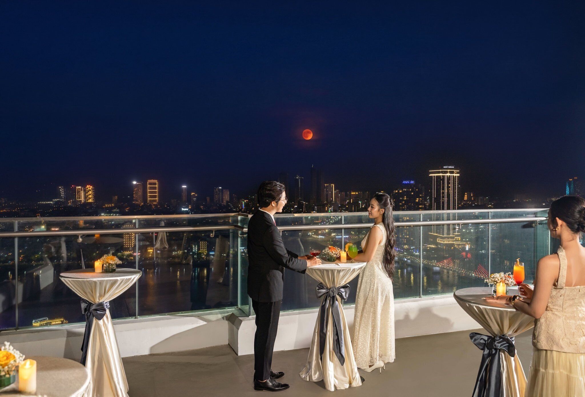 Une terrasse sur le toit, avec des tables dressées pour un mariage, offrant une vue sur le paysage urbain illuminé la nuit.