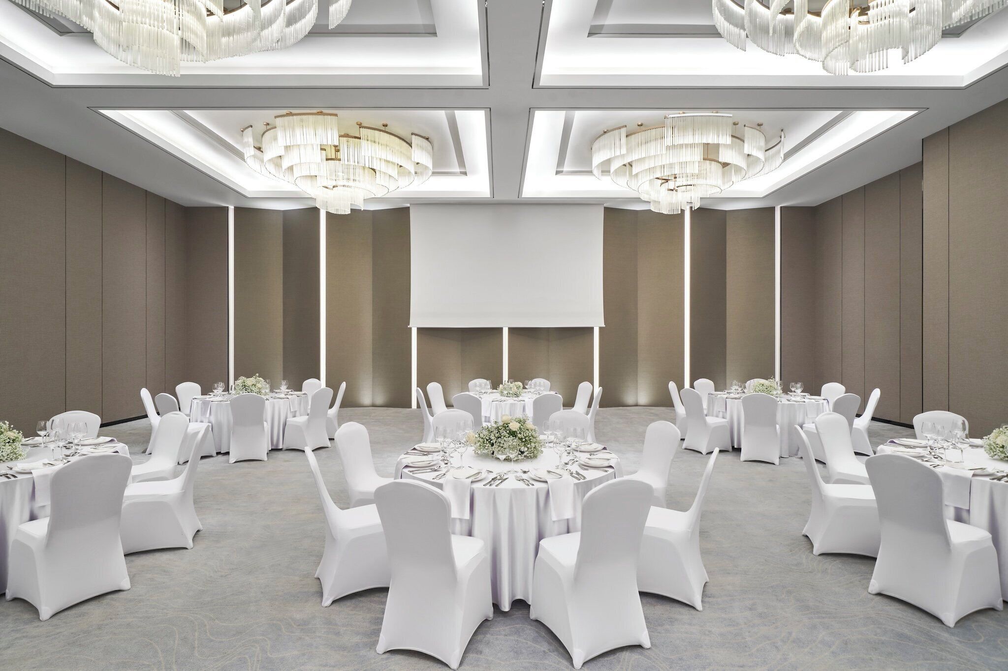 A spacious banquet hall with round tables, white chairs, and chandeliers.