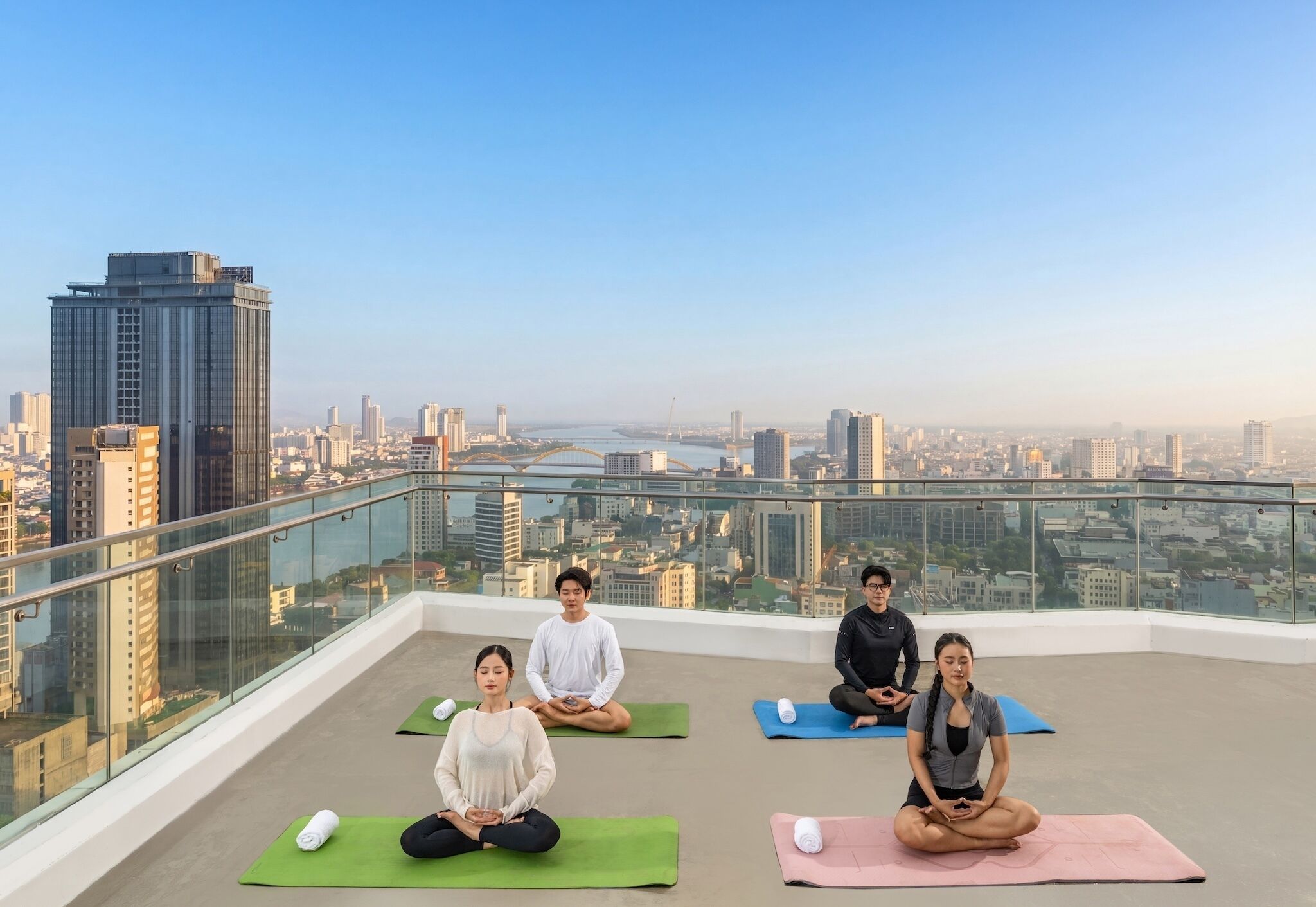 A rooftop with four individuals meditating on yoga mats, overlooking a cityscape with high-rise buildings and a river.