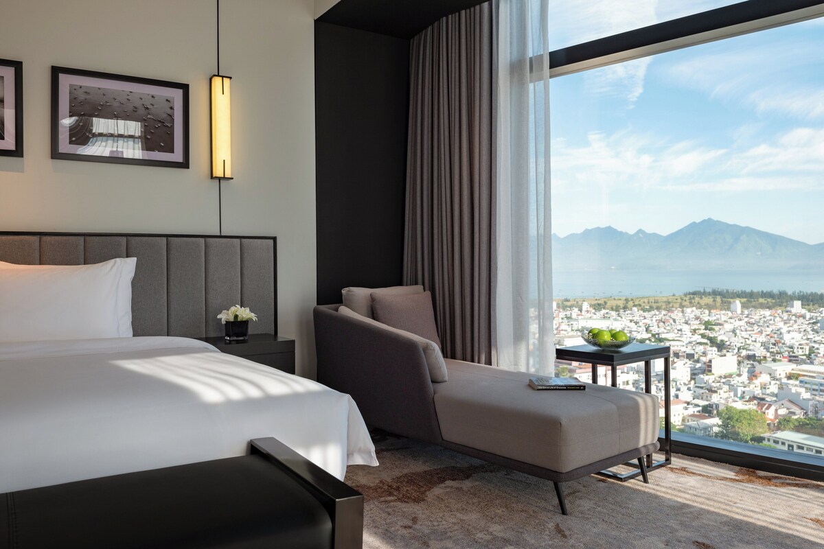Una habitación de hotel moderna con cama, zona de estar, mesita de noche y un amplio ventanal con vistas a la ciudad y la montaña.