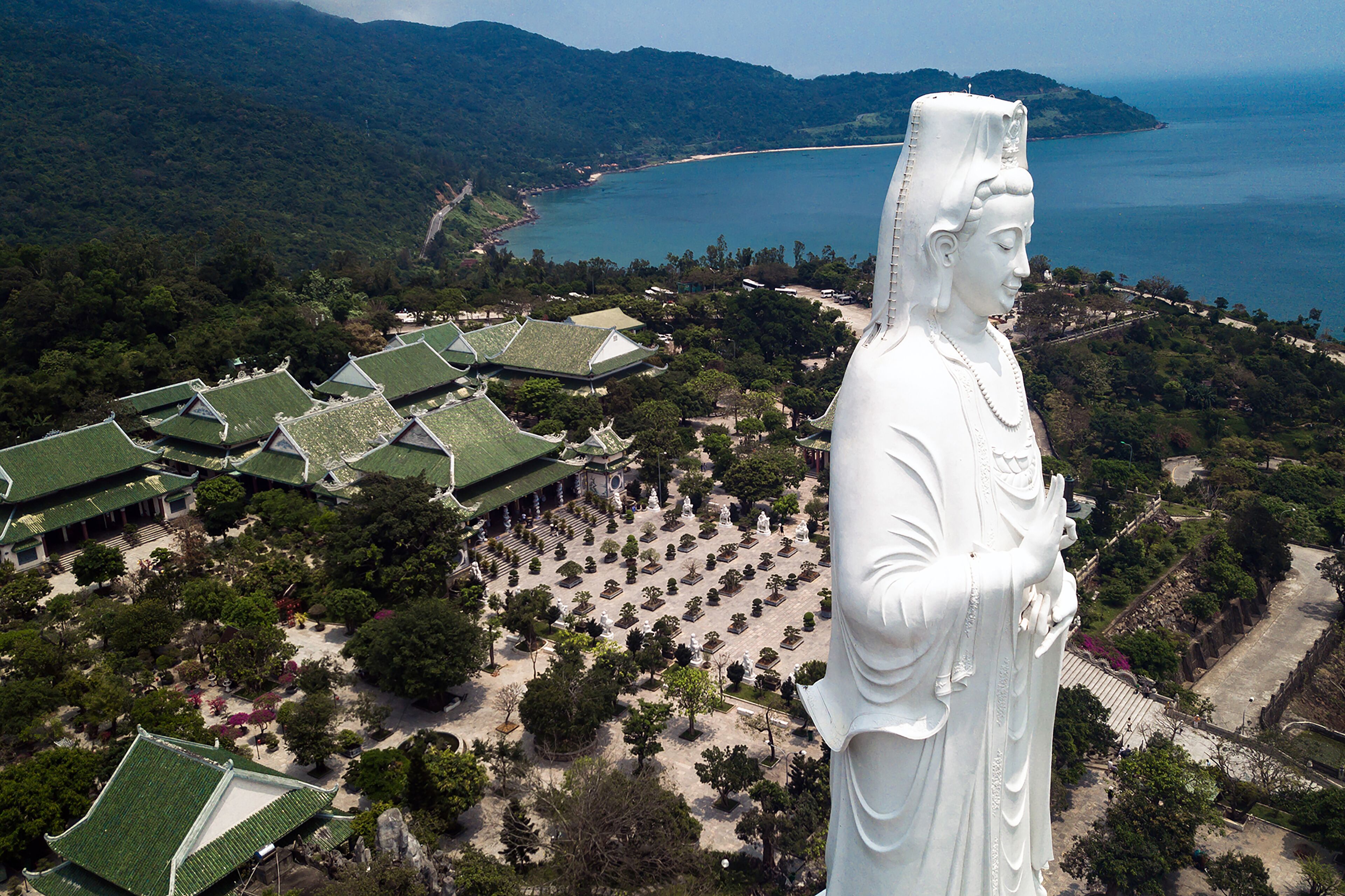 A large white statue overlooking a traditional building complex with green roofs, surrounded by a landscaped garden and a coastal view.