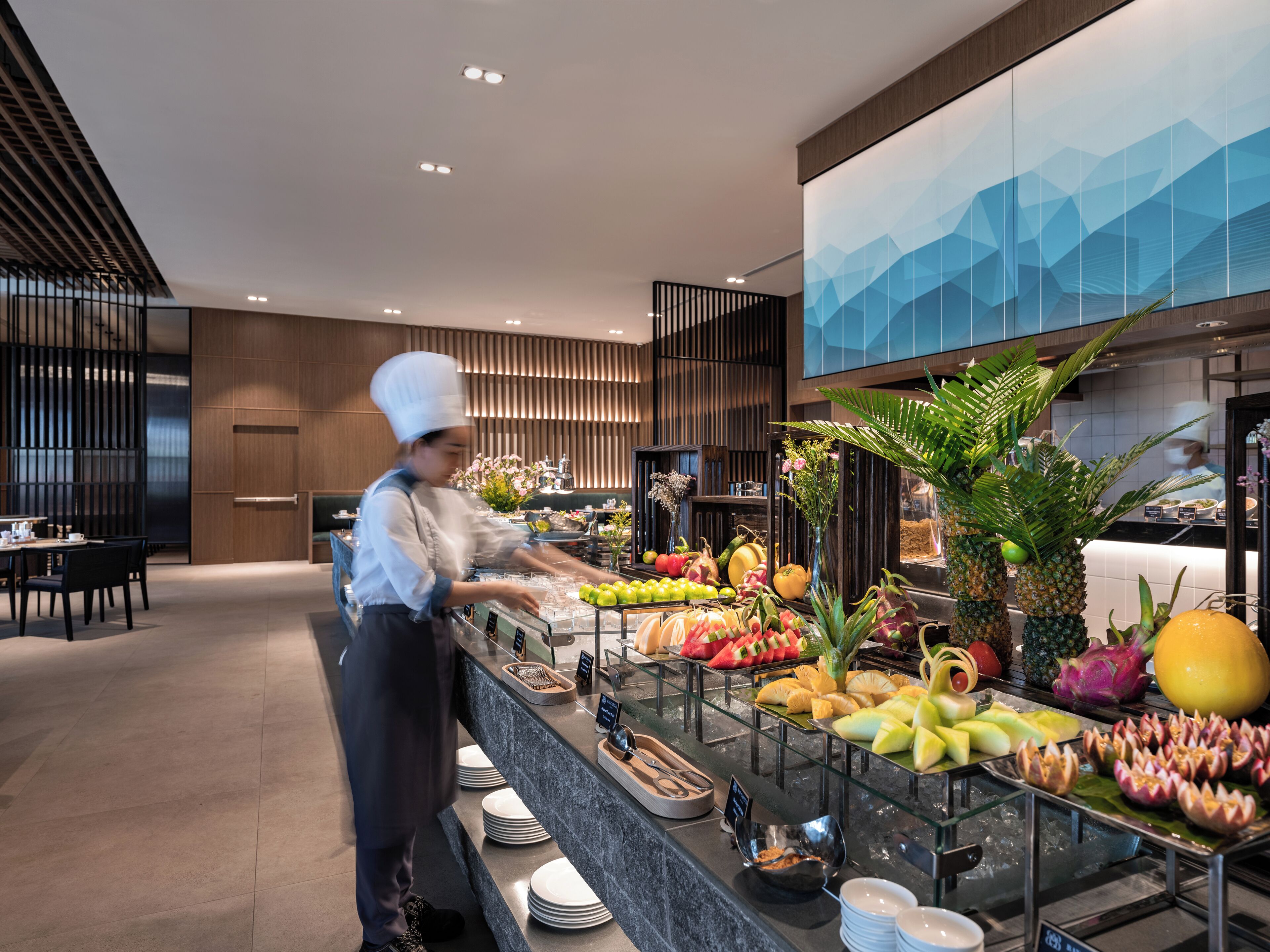 A buffet area with a variety of fruits and vegetables, a chef preparing food, and a modern interior design.