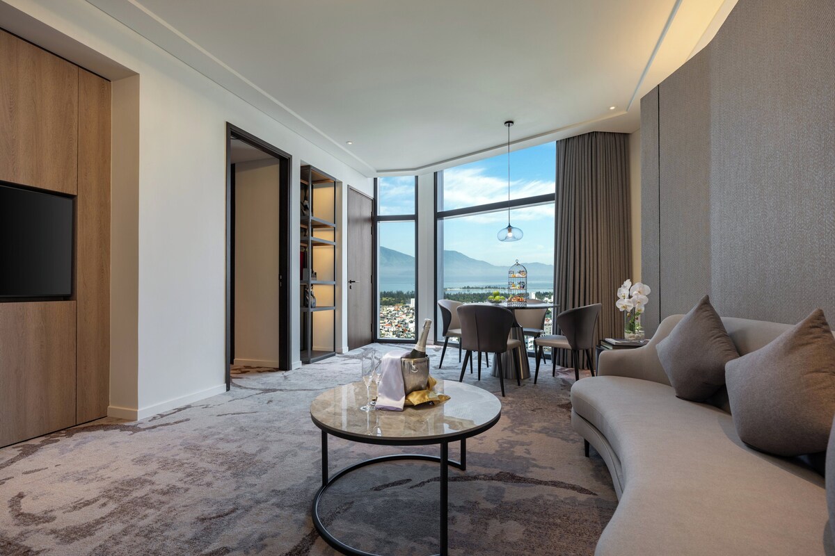 Una habitación de hotel moderna con un ventanal grande que ofrece vistas panorámicas, un espacio de descanso acogedor con sofá y sillones, y un televisor de pantalla plana montado en la pared.