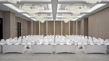 A conference room with rows of white chairs and tables, large chandeliers, and a high ceiling with skylights.