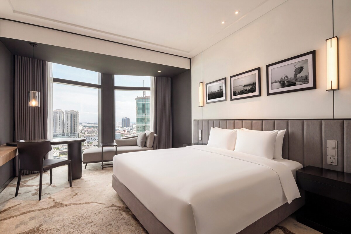 A modern hotel room with a large bed, a desk, and a view of the cityscape.