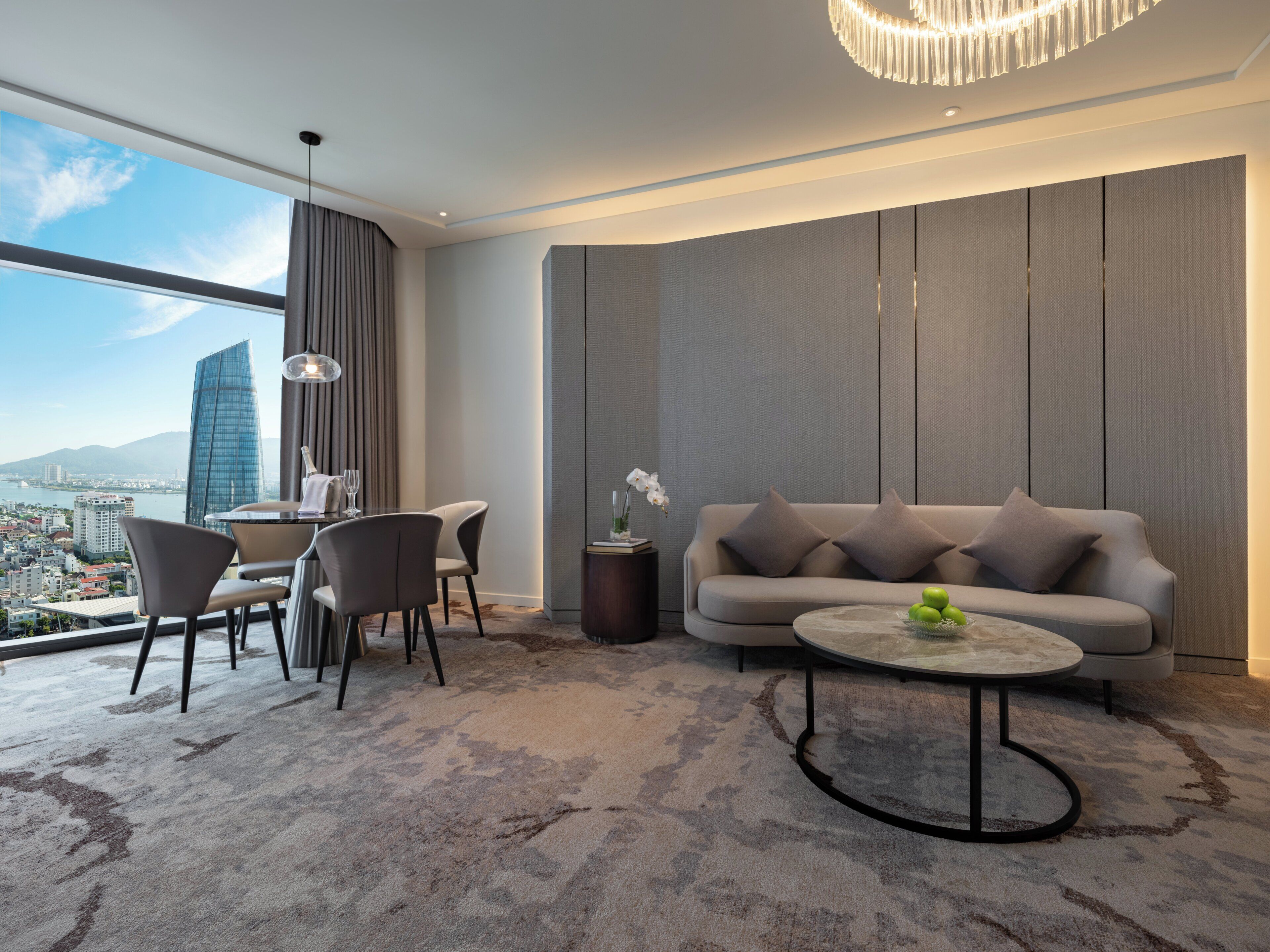 Ein modernes Hotelzimmer mit großem Fenster und Blick auf die Stadt, einer Sitzecke, einem Essbereich mit Stühlen und einem runden Couchtisch.