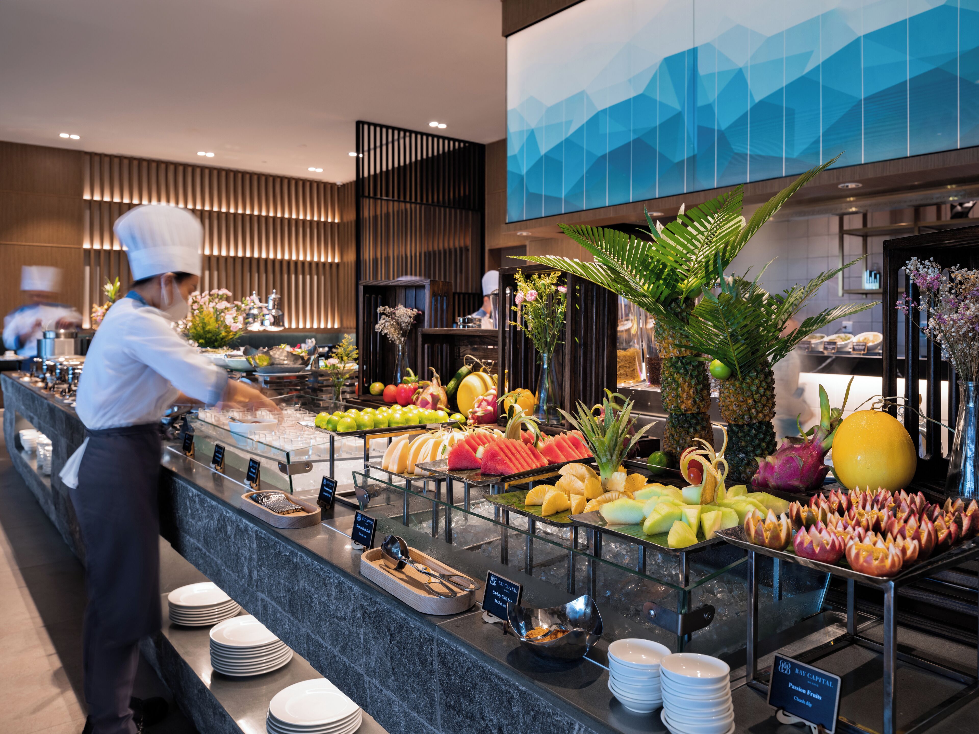 A buffet area with a variety of fruits and vegetables, a chef preparing food, and a modern interior design.