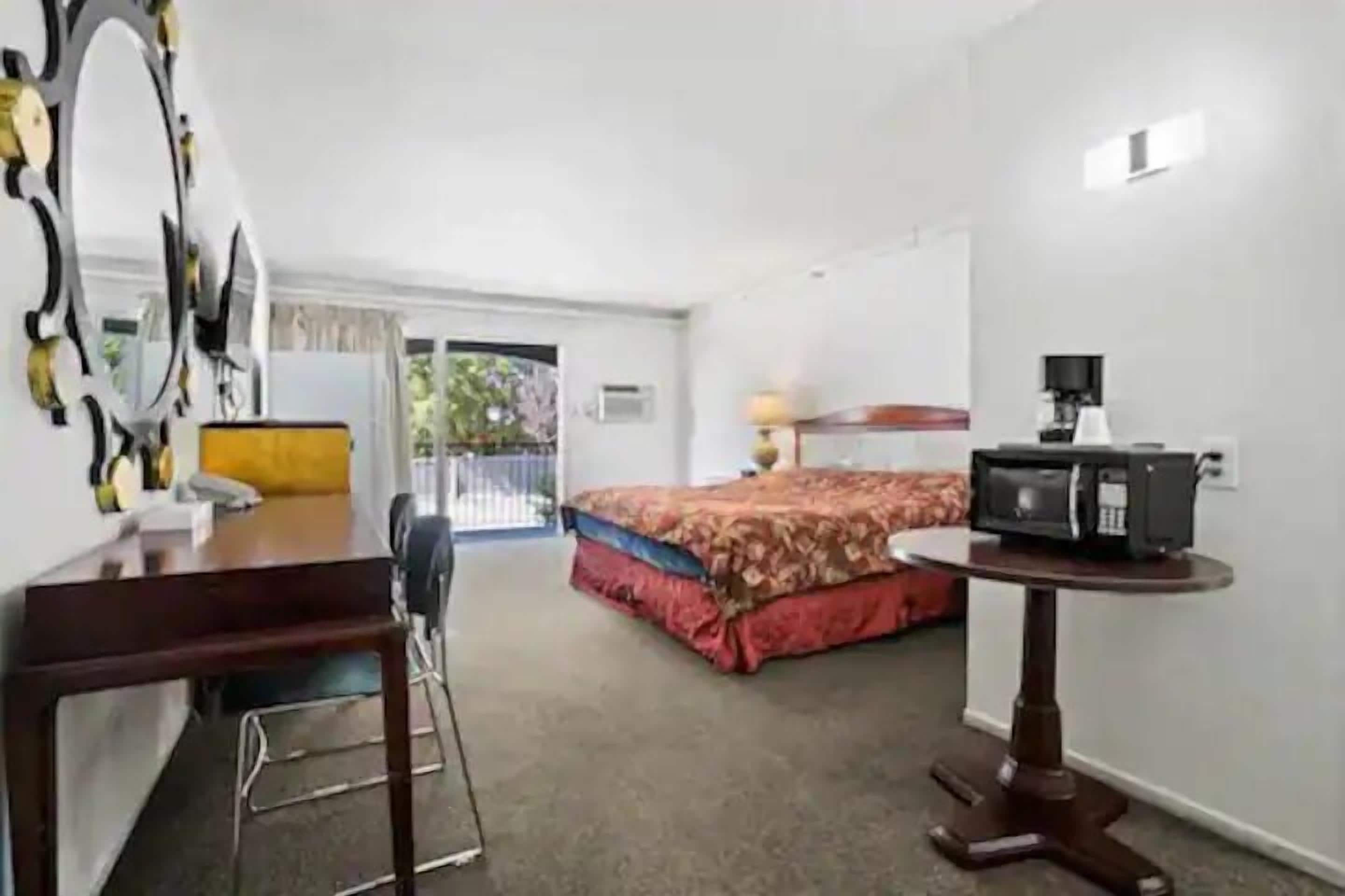 A hotel room with a bed, a desk with a chair, a small table with a lamp, and a microwave on a stand.