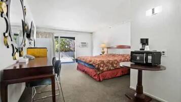A hotel room with a bed, a desk with a chair, a small table with a lamp, and a microwave on a stand.