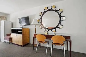 Habitación de hotel con escritorio de madera, dos sillas, televisor sobre un mueble bajo y un espejo decorativo en la pared.