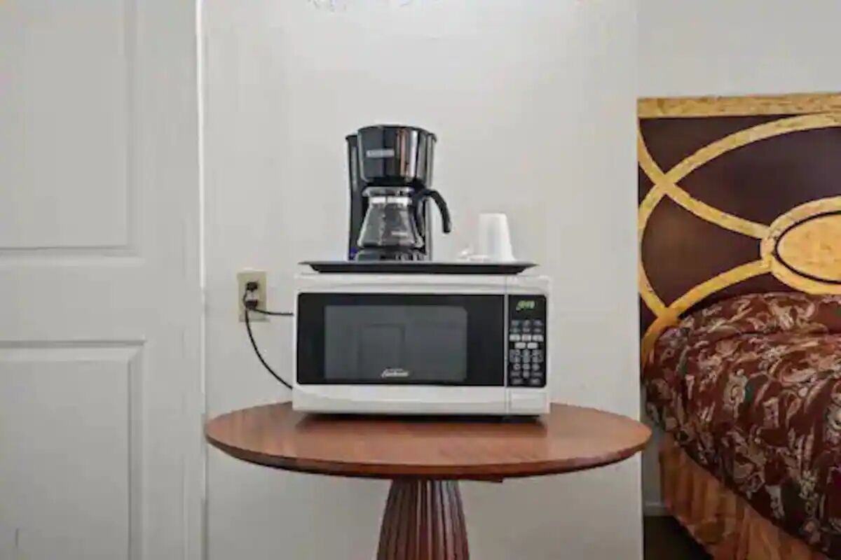 A white microwave on a small wooden table in a room with a patterned bedspread.