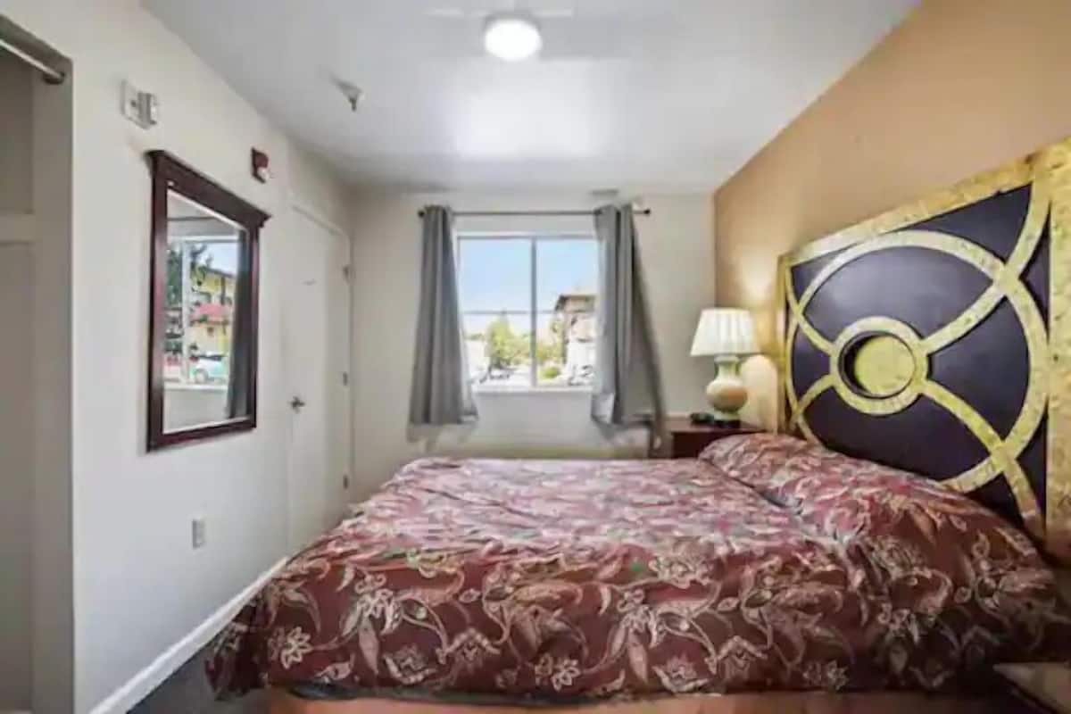 Un dormitorio con cama, ventana con cortinas, lámpara de pared y cabecera decorativa.