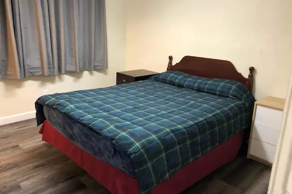 Una cama individual con cubrecama escocés y cabecera de madera en una habitación con suelo de madera y una mesita de noche pequeña.