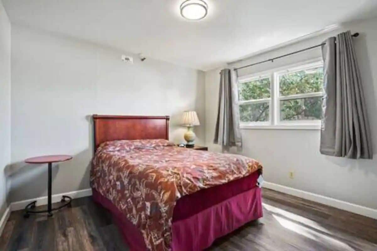 Un dormitorio con una cama de madera, una mesita redonda pequeña, una lámpara y una ventana con cortinas.