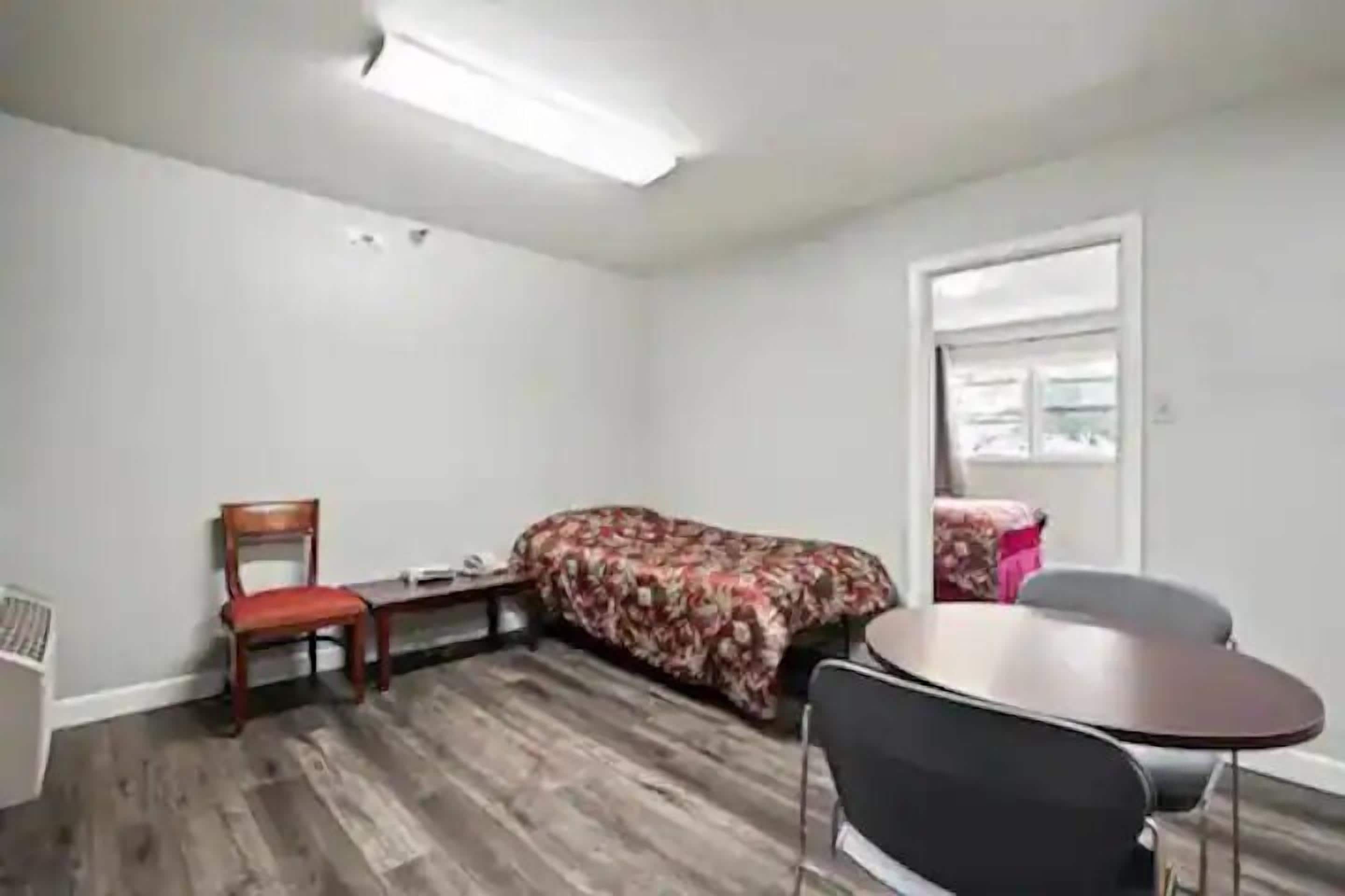 A small, sparsely furnished room with a wooden floor, a single bed with a floral bedspread, a chair, a small table, and a window with a view of the outside.