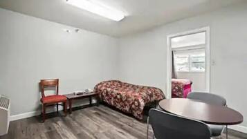 A small, sparsely furnished room with a wooden floor, a single bed with a floral bedspread, a chair, a small table, and a window with a view of the outside.