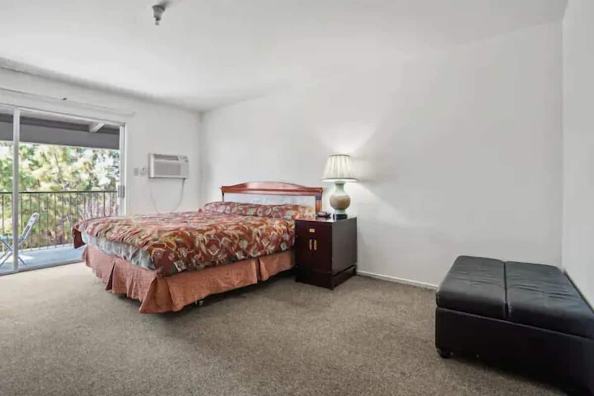Un dormitorio con cama, mesita de noche, lámpara, banca y puerta corrediza de vidrio a un balcón.