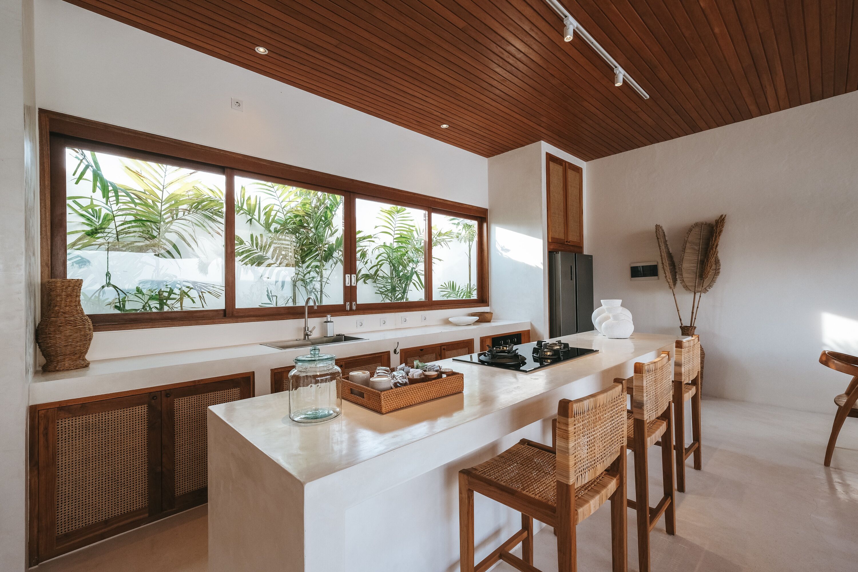 A modern kitchen with a large island, wooden ceiling, and large windows overlooking greenery.