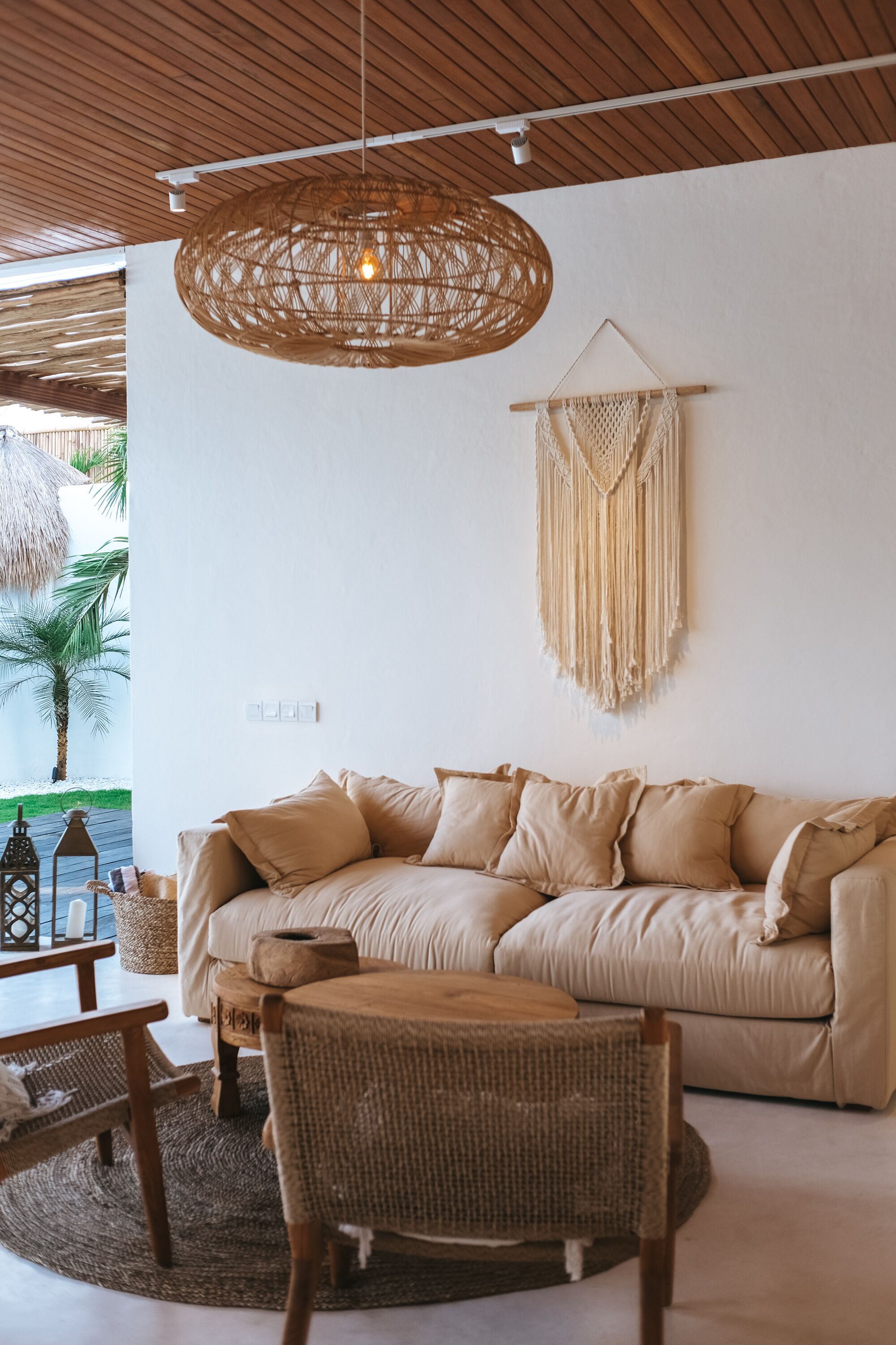 A cozy living room with a beige sofa, a wicker chair, a round wooden table, a macrame wall hanging, and a woven pendant light.