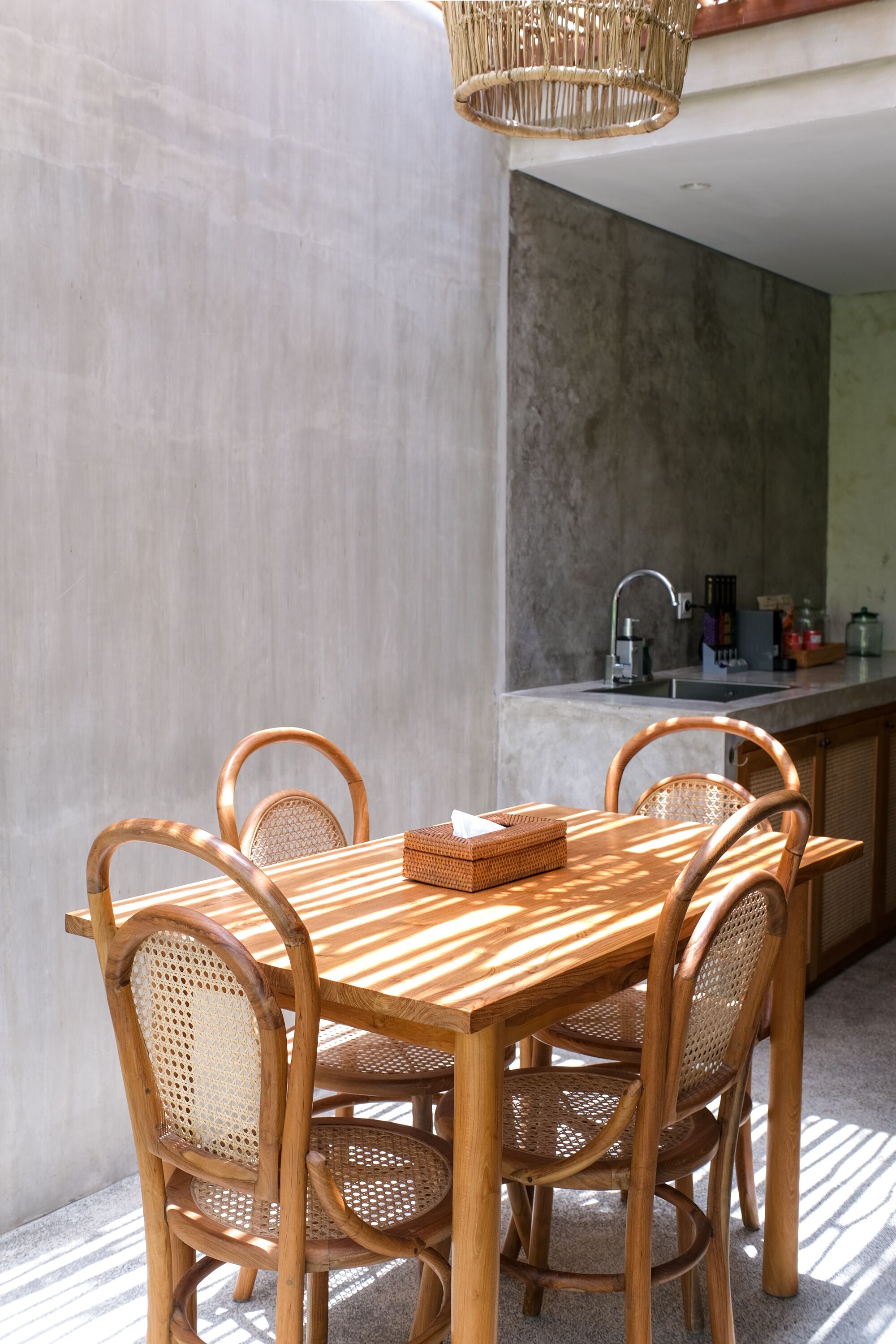 Una mesa de madera con cuatro sillas, una cesta tejida y una cocina con pileta y alacenas.