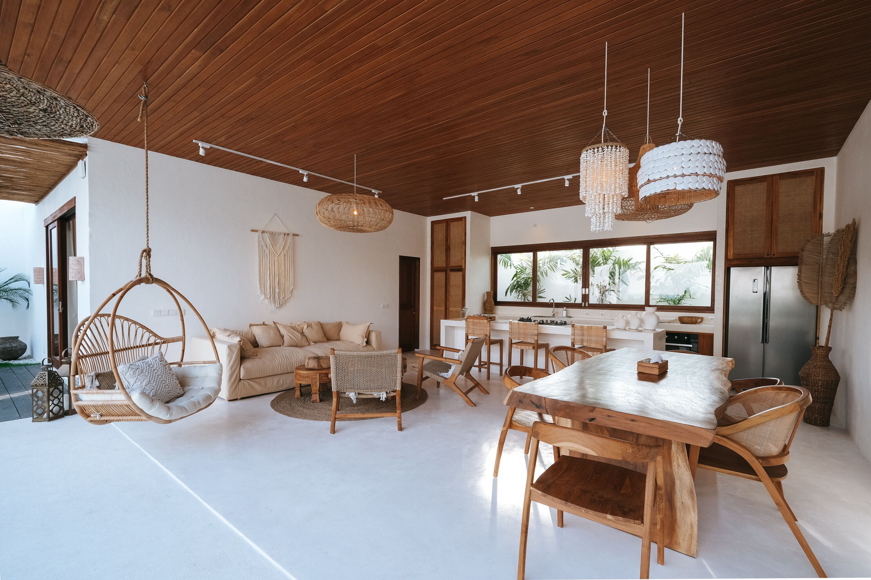 A spacious living area with a wooden ceiling, hanging wicker chairs, a dining table with chairs, and a kitchen area with a refrigerator.