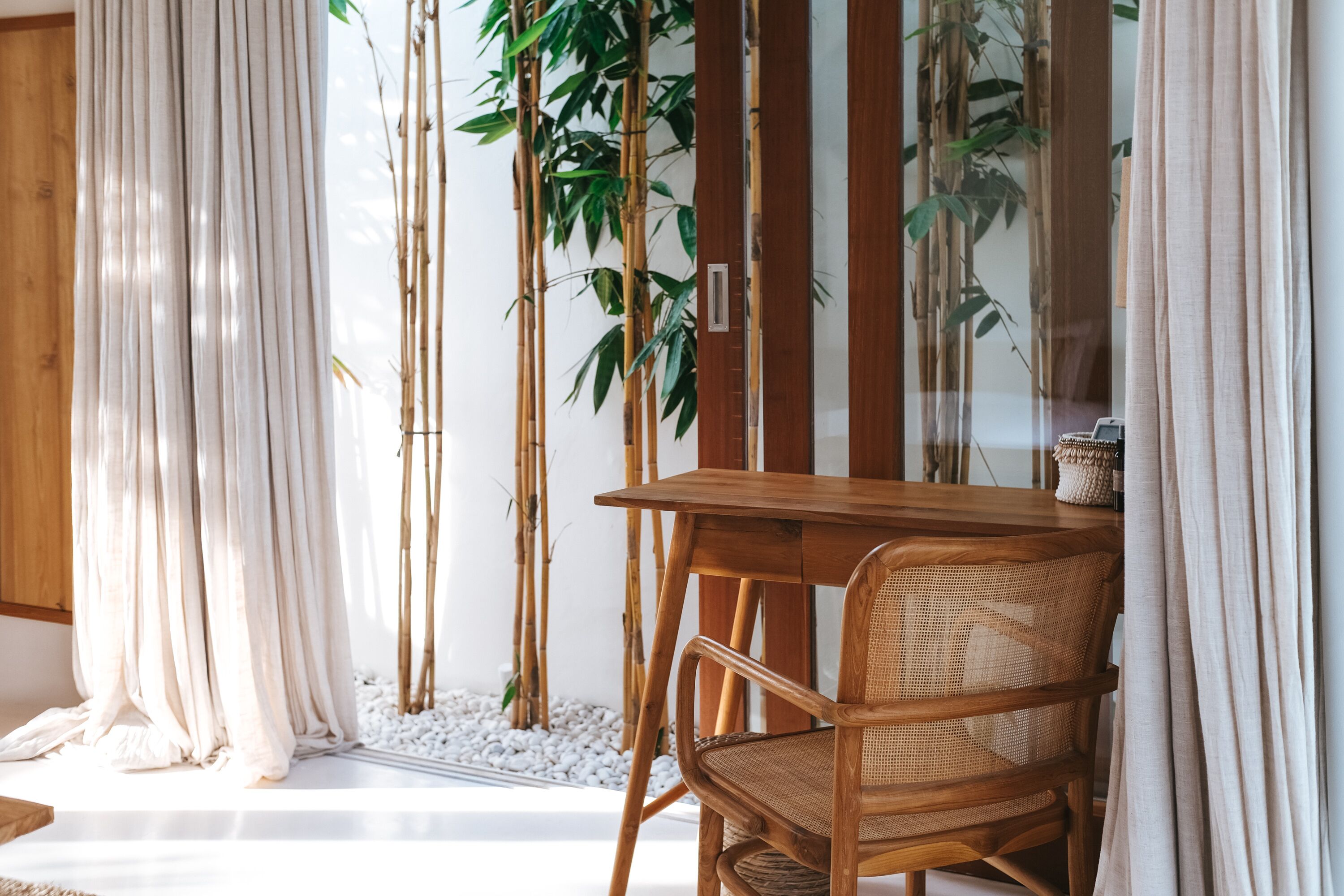Une pièce avec une table en bois et une chaise en osier, une plante en bambou et des rideaux blancs.