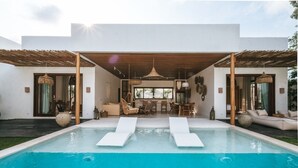 Un área de piscina moderna con dos sillones de descanso, un comedor con mesa y sillas, y un gran ventanal con vista a la piscina.