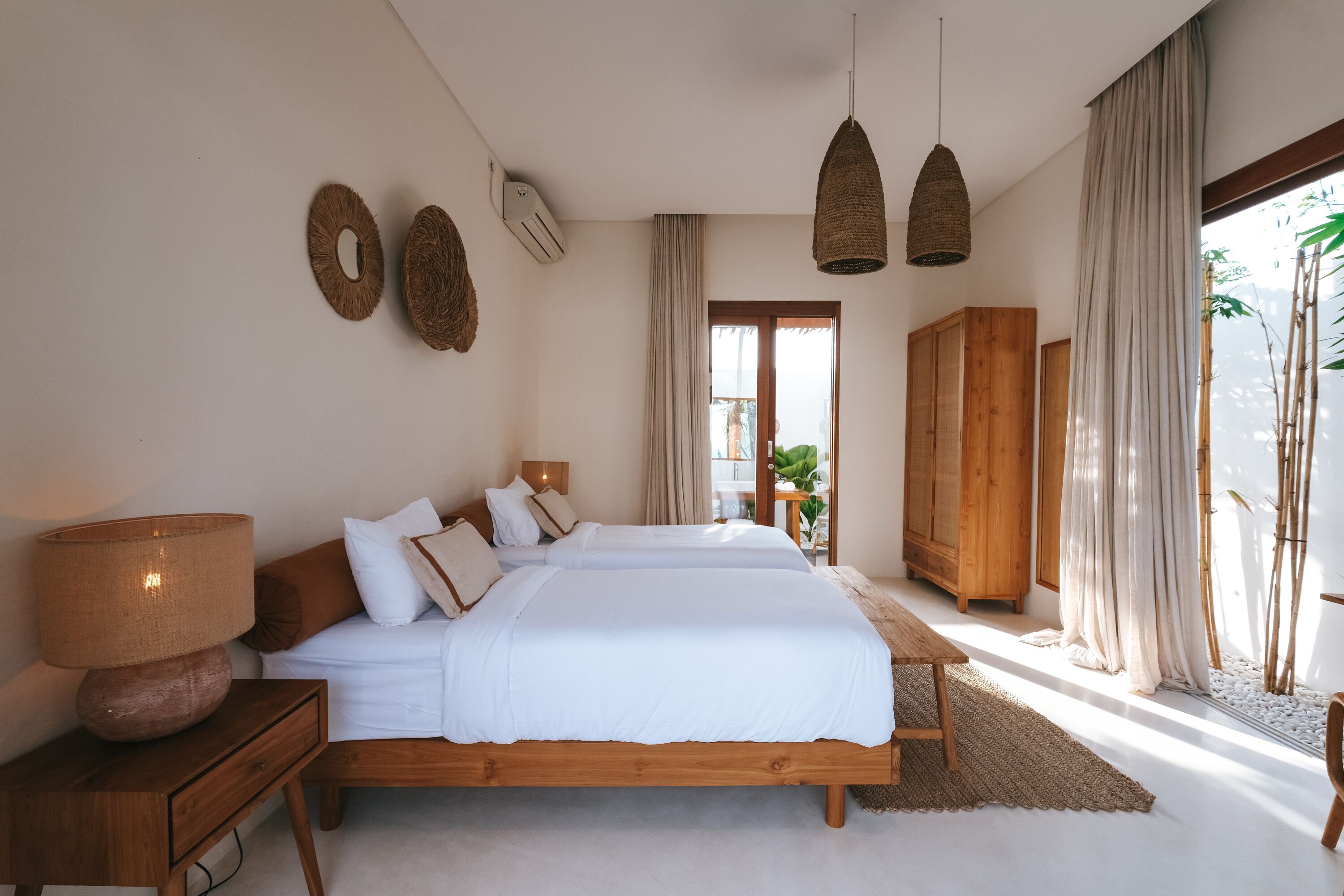 A bedroom with two beds, a wooden wardrobe, a bedside table with a lamp, and a large window with curtains.