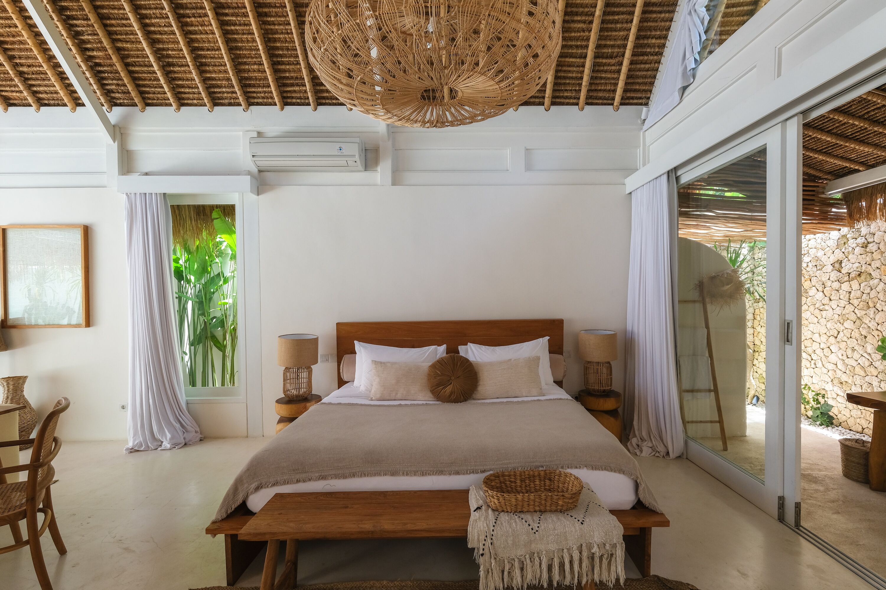 Une chambre avec un grand lit, un banc en bois, un panier en osier et une vue sur un mur de pierre et la verdure environnante.