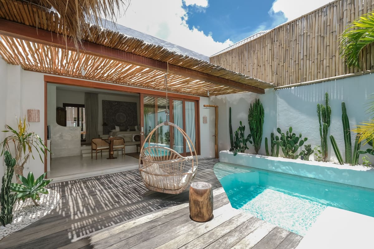 A poolside area with a wicker chair, a wooden stool, and a variety of cacti.