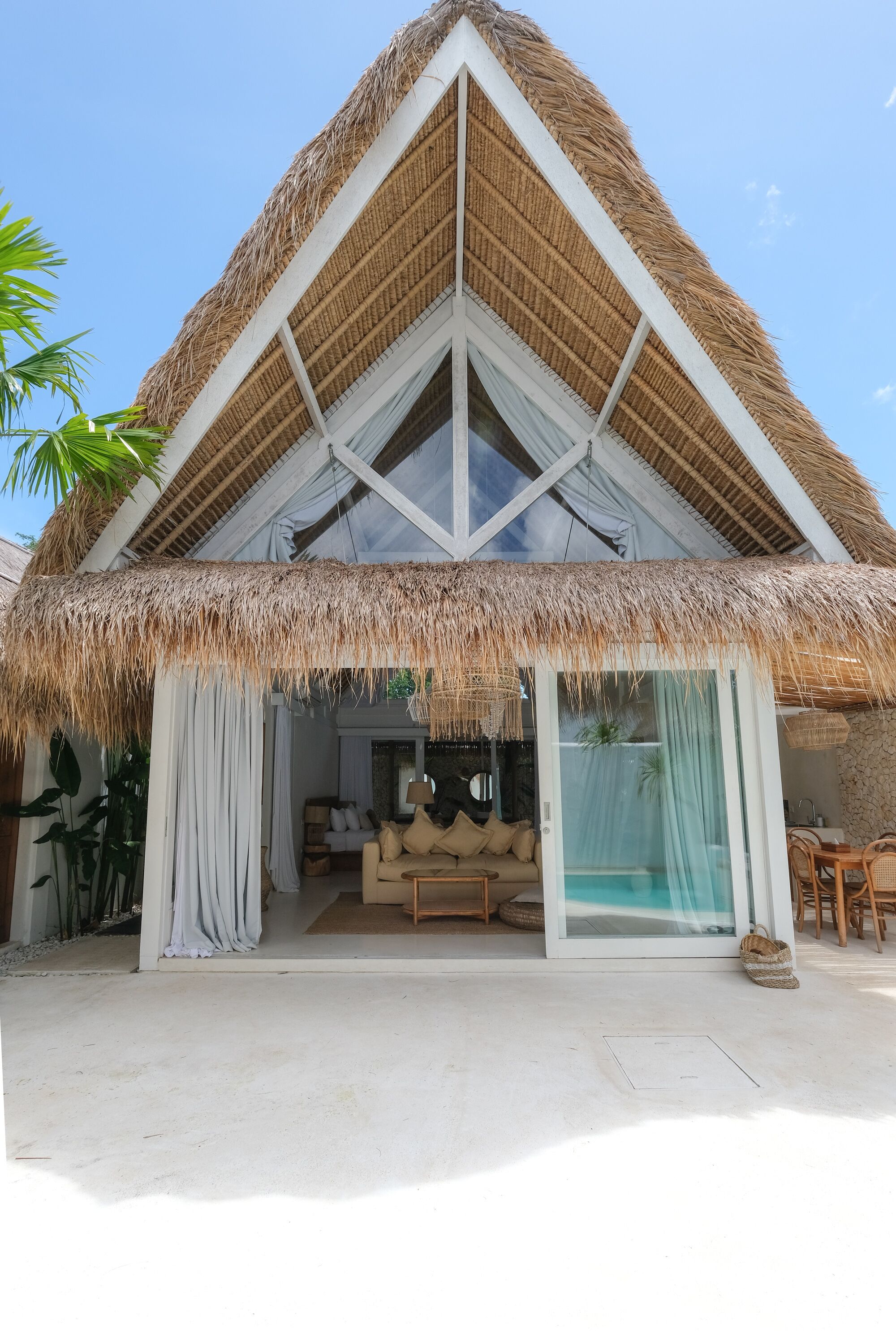 A thatched-roof villa with a glass-enclosed living area, a pool, and a dining area.