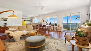 A spacious living room with a sofa, ottoman, and dining area. A kitchen is visible in the background, and large windows offer a view of the ocean.