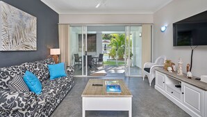 Ein modernes Wohnzimmer mit einem gemusterten Sofa, einem Couchtisch, einem Fernseher und einer Schiebetür zu einem Außenbereich.