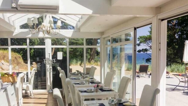 Un comedor iluminado por el sol, con mobiliario blanco, una araña y vistas al exterior.
