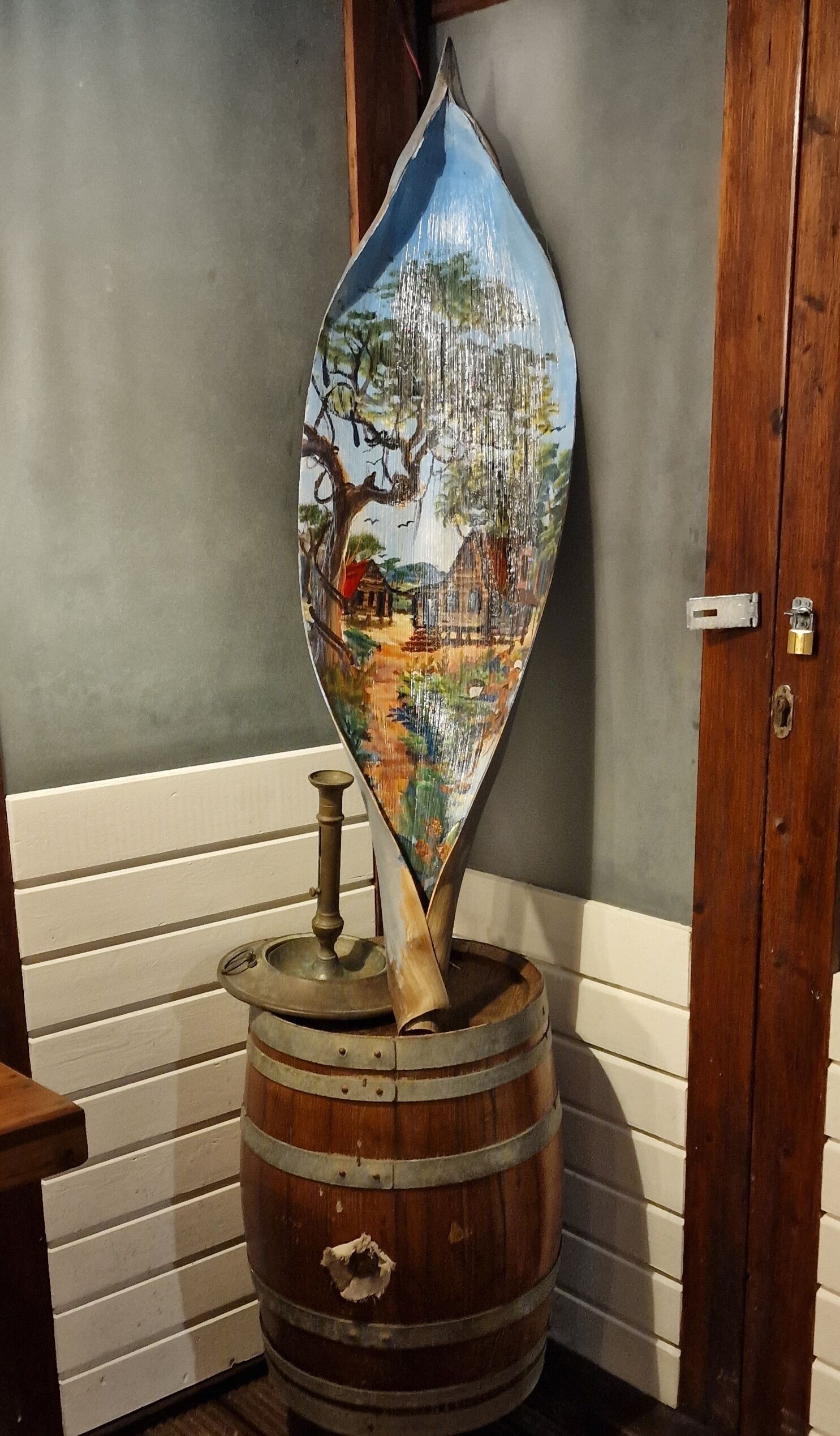 A wooden barrel with a painted depiction of a tree and landscape, placed against a wall with a door.