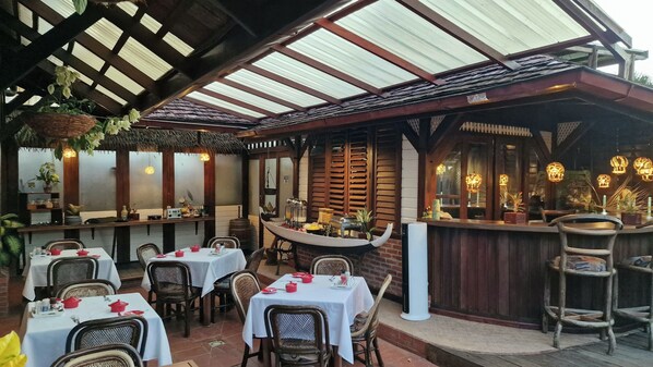 An outdoor restaurant with tables and chairs, a bar area, and a thatched roof. - Greenheart Boutique Hotel (Paramaribo)