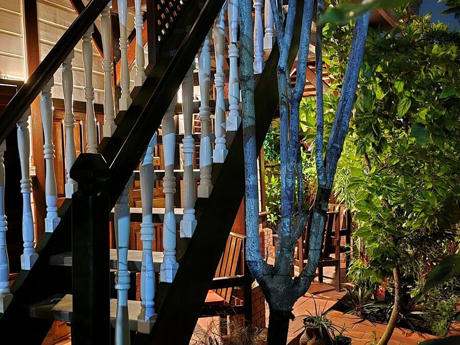 Una escalera de madera con barandillas blancas que conduce a un espacio al aire libre techado, con piso de ladrillo y plantas en macetas.
