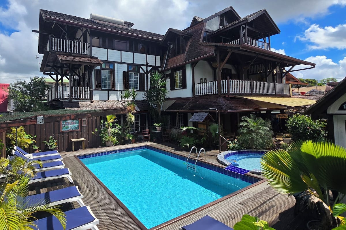 A two-story building with a swimming pool, lounge chairs, and tropical plants.
