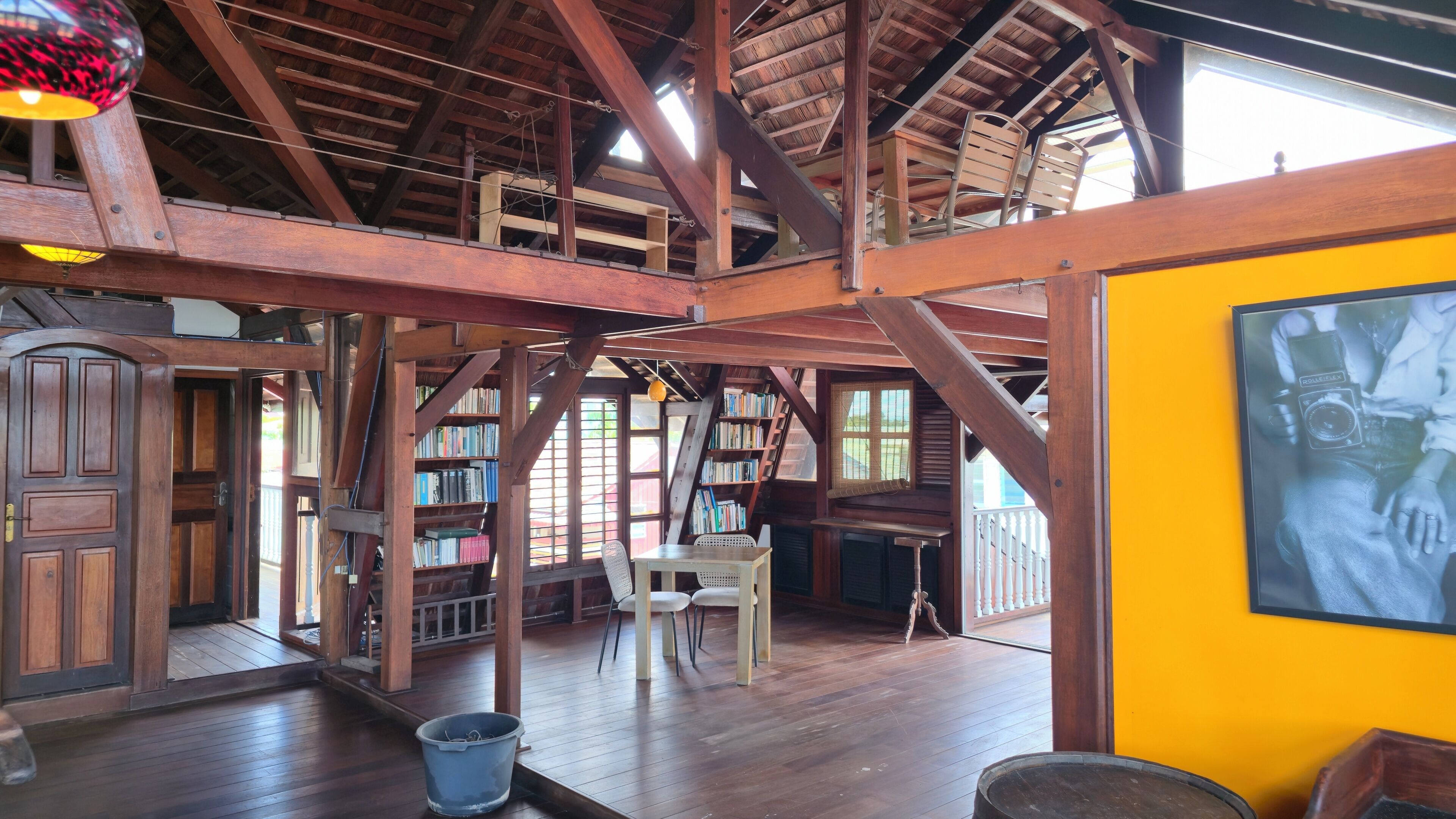 Una habitación amplia con techo alto, vigas de madera, una zona de comedor con mesa y sillas, y una estantería llena de libros.