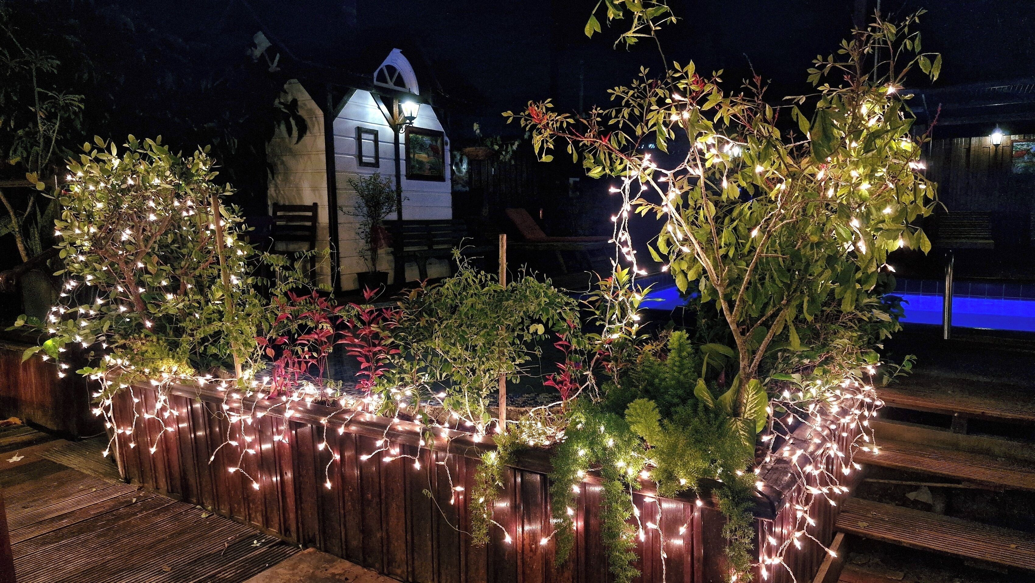 Una terraza de madera con luces cálidas y plantas, un edificio blanco con techo a dos aguas al fondo y una zona de piscina iluminada en azul.