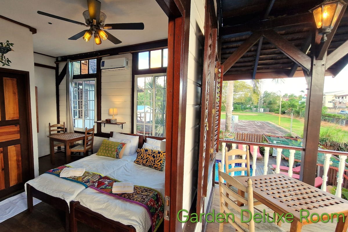 A bedroom with a bed, a chair, a table, and a ceiling fan. There is a window with curtains, a door, and a view of a garden and pool outside.
