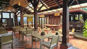 An outdoor dining area with wooden tables and chairs, a brick fireplace, and a thatched roof. - Greenheart Boutique Hotel (Paramaribo)
